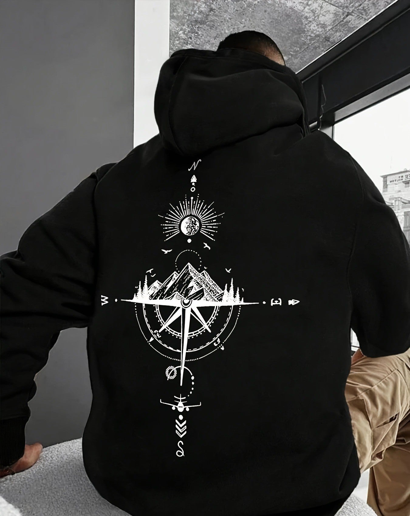 Person wearing a black hoodie with a white compass design on the back, standing indoors.