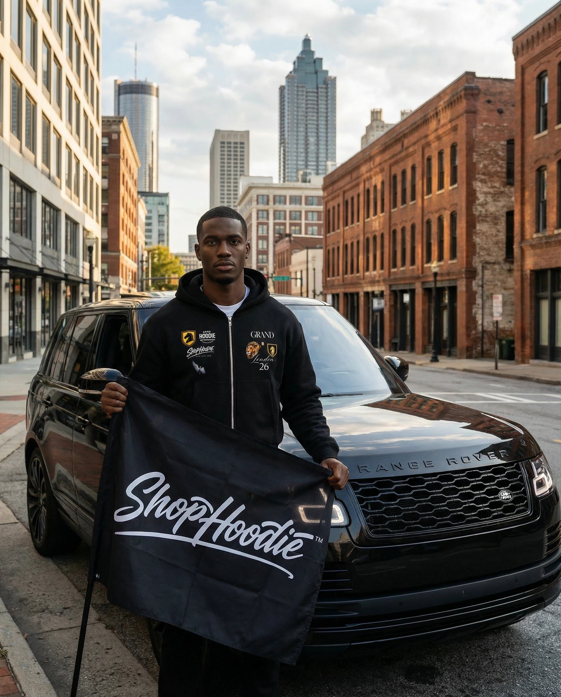 Man holding a 'ShopHoodie' jacket in front of a black SUV with cityscape background