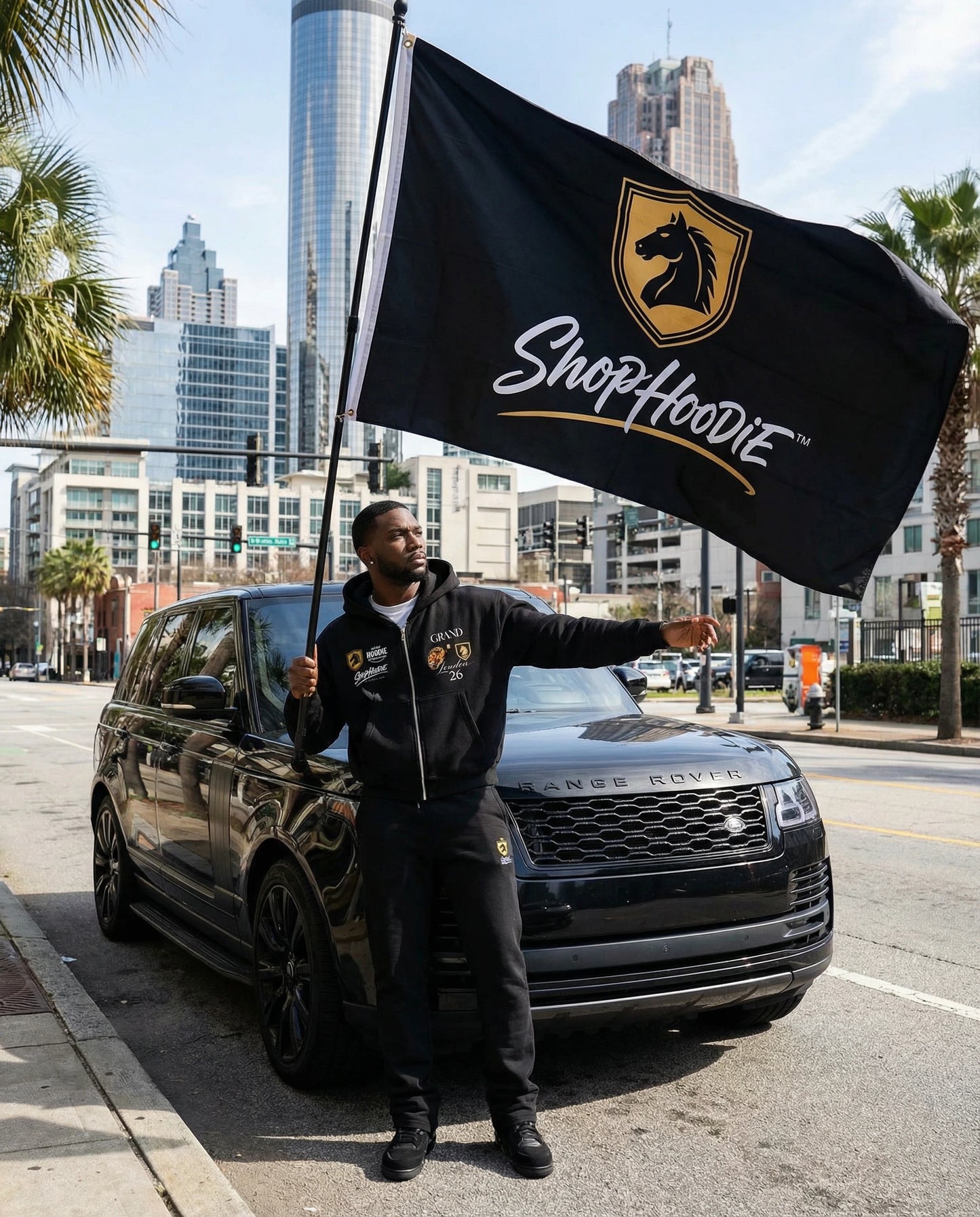 Person holding a ShopHoodie flag next to a black SUV on a city street.