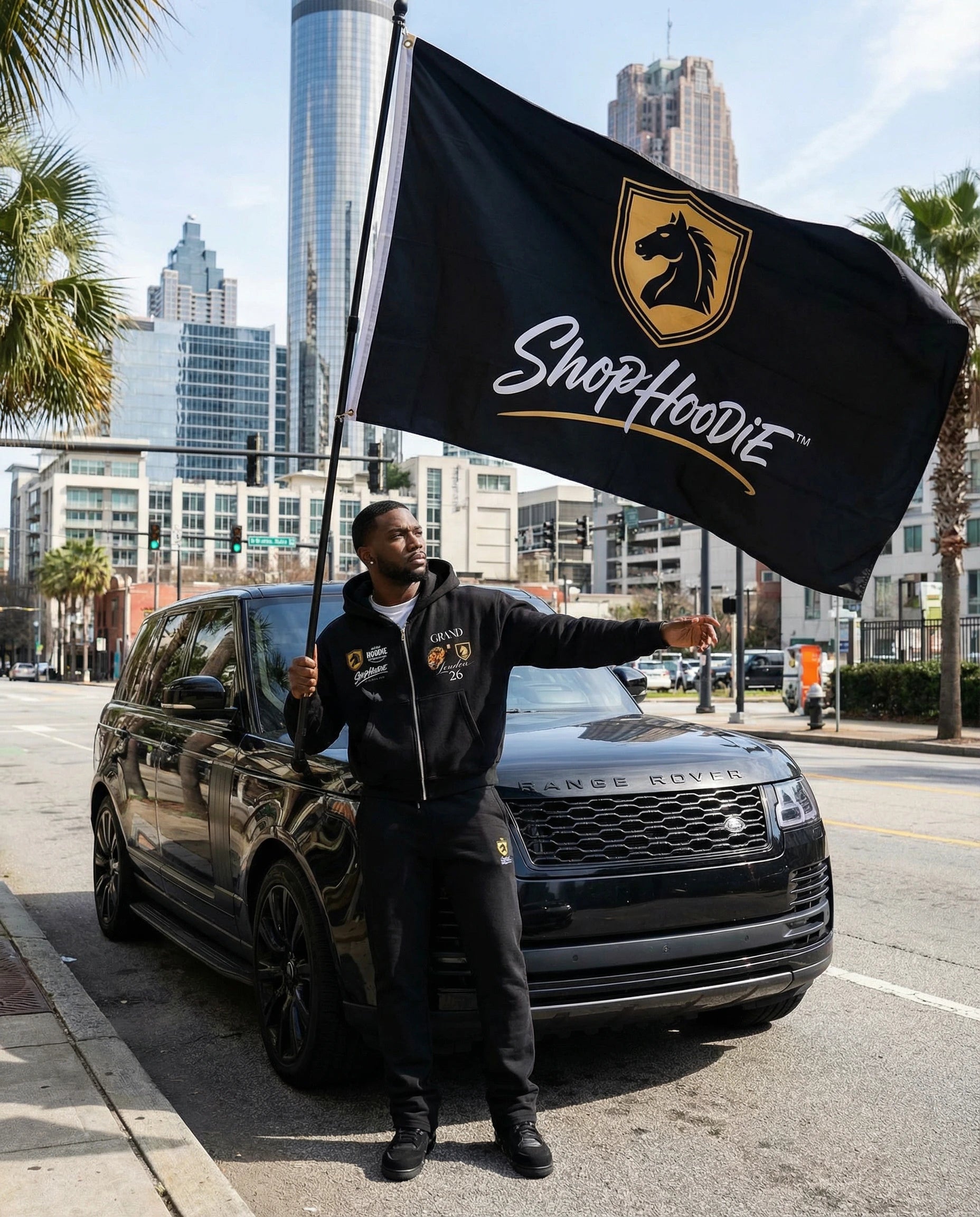 Person holding a ShopHoodie flag next to a black SUV on a city street.