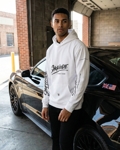 Man wearing a white hoodie with branding, standing next to a black car in an urban setting.