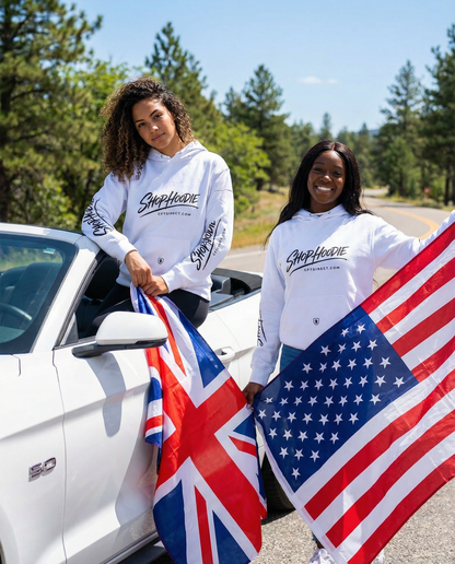 Two people wearing 'Sheep Herder' sweatshirts holding British and American flags by a white truck.