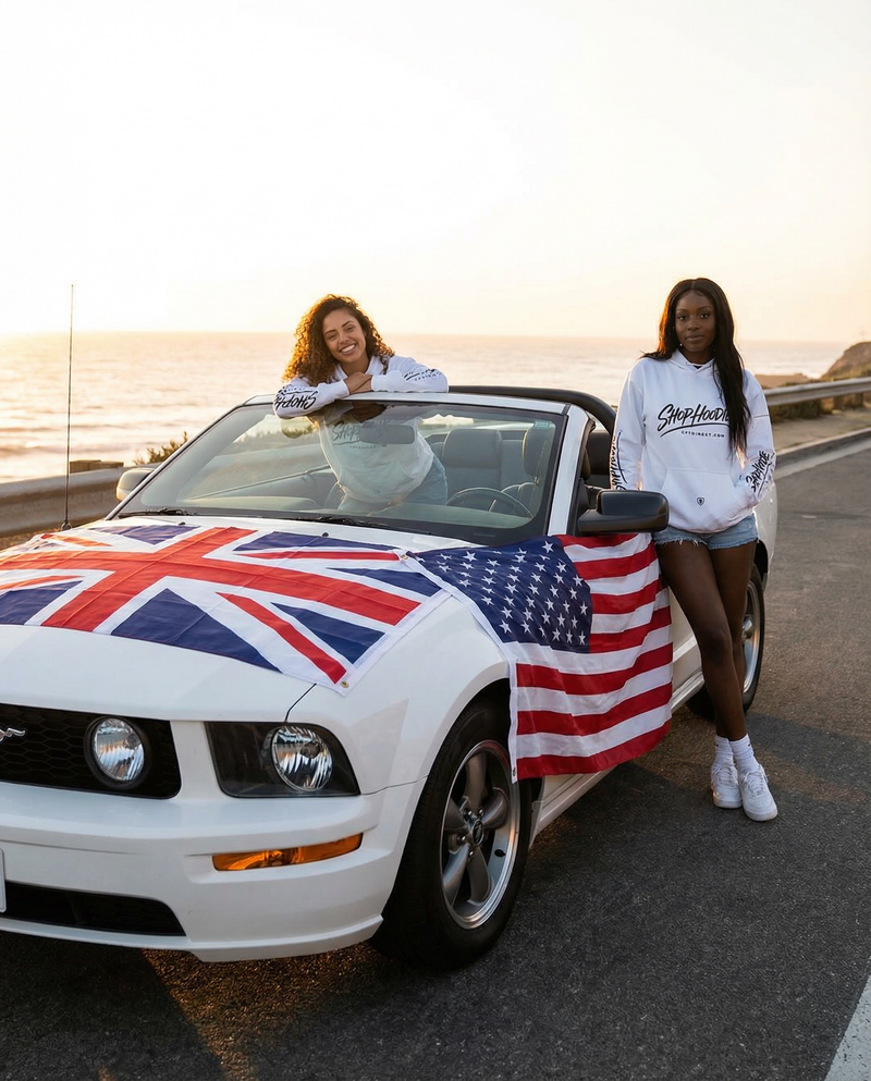 Two women standing next to a convertible car with a British and American flag draped over it, on a scenic road.