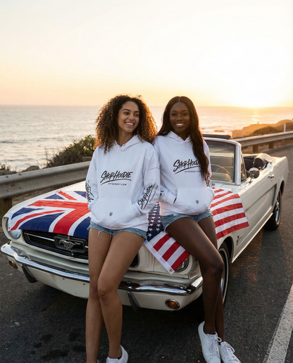 Two women in white hoodies standing next to a classic car with a Union Jack and American flag design, by the ocean.