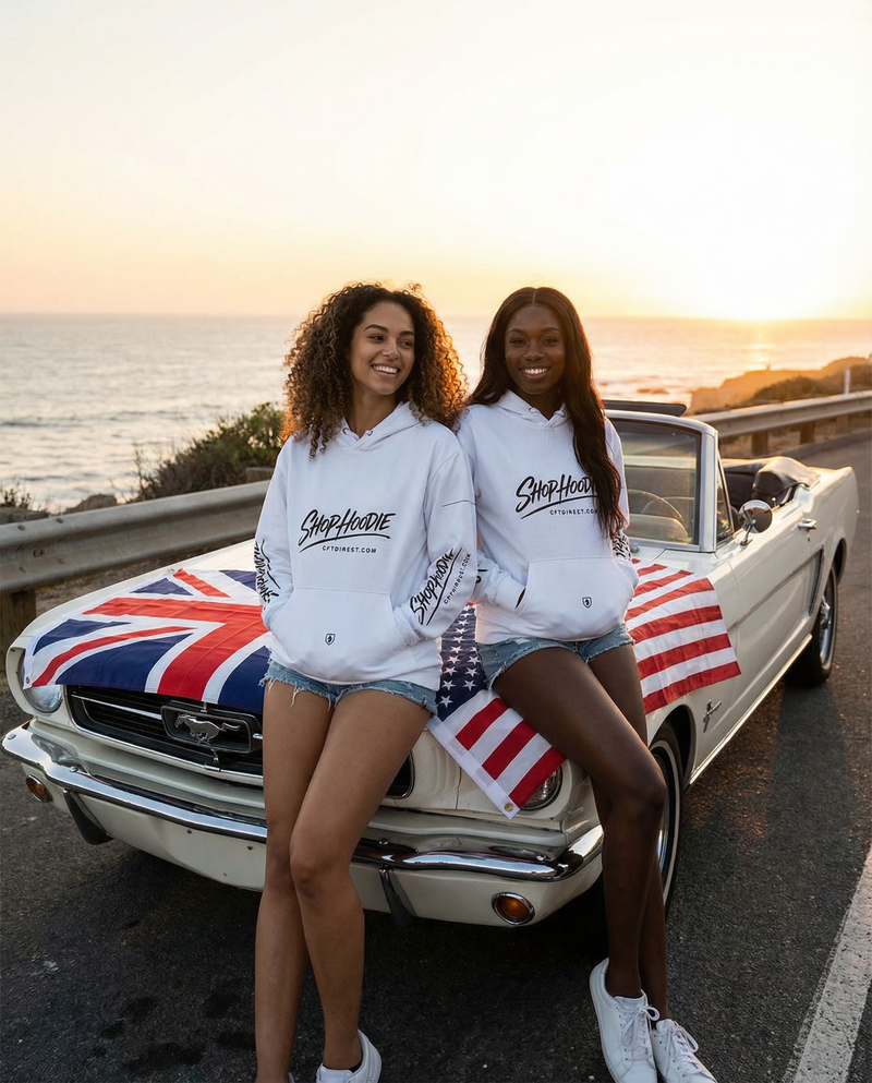 Two women in white hoodies standing next to a classic car with a Union Jack and American flag design, by the ocean.