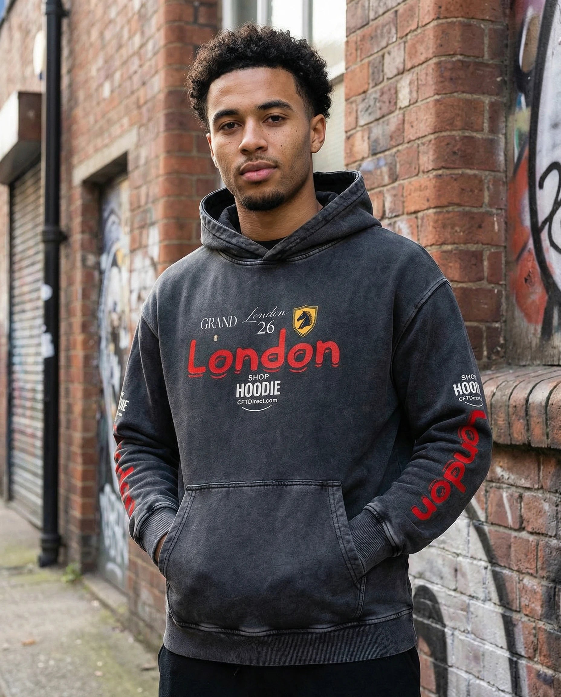 Person wearing a hoodie with 'Grand London 26' text and logo, standing against a brick wall with graffiti.
