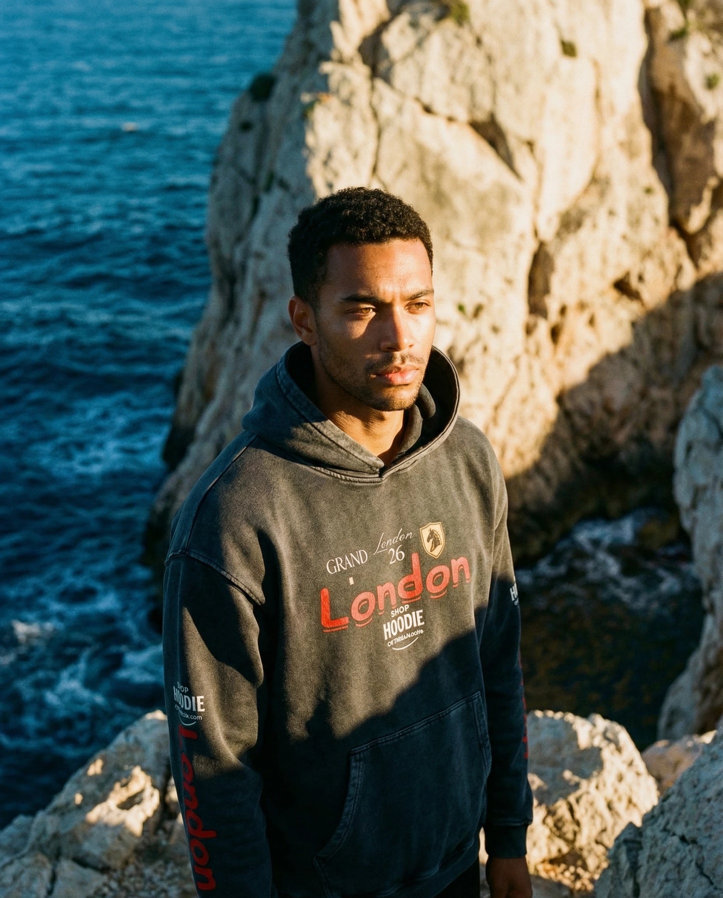 Man wearing a hoodie with text standing on rocks by the sea
