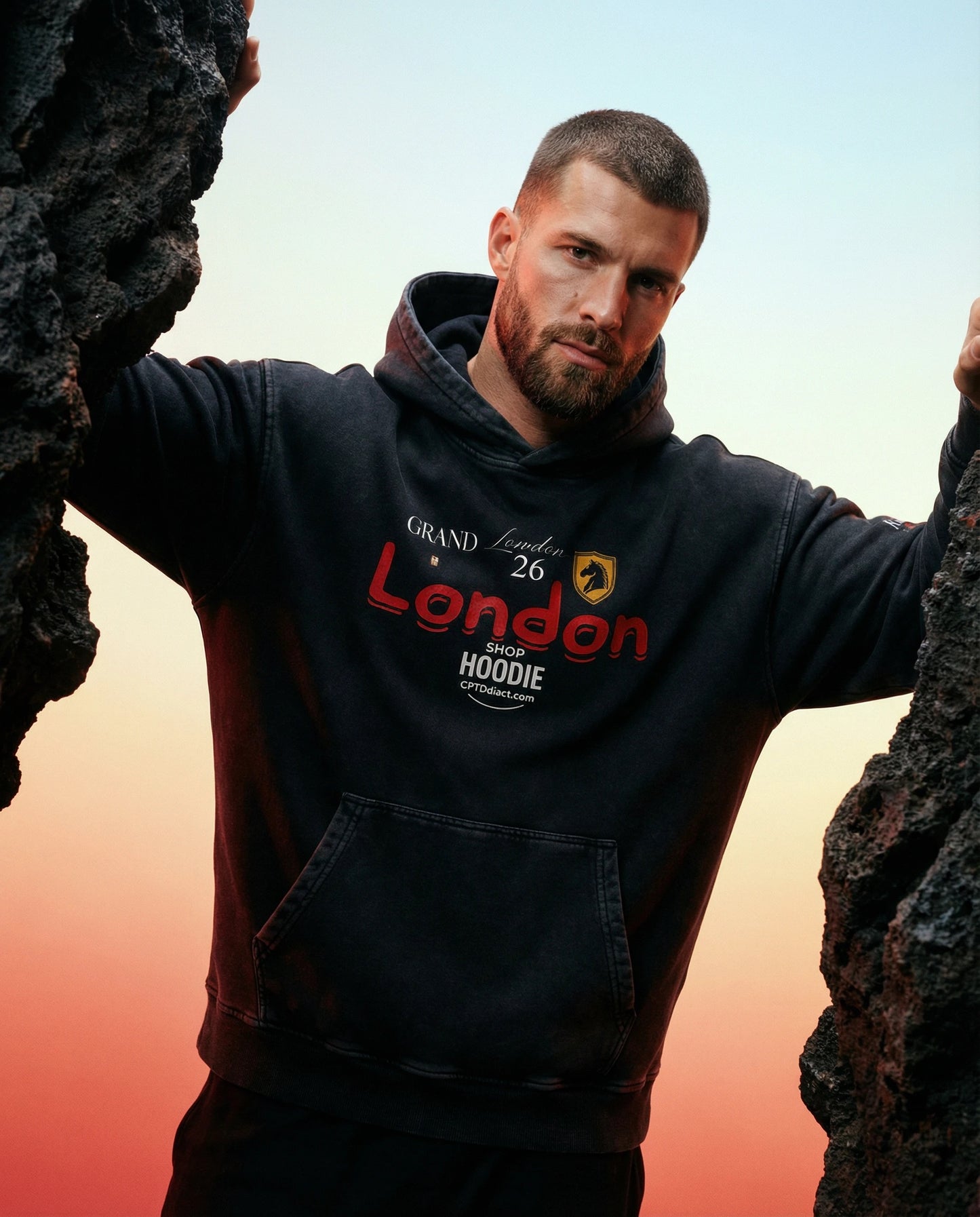 Man wearing a hoodie with 'Grand London' branding, standing between two rocks against a sunset sky.