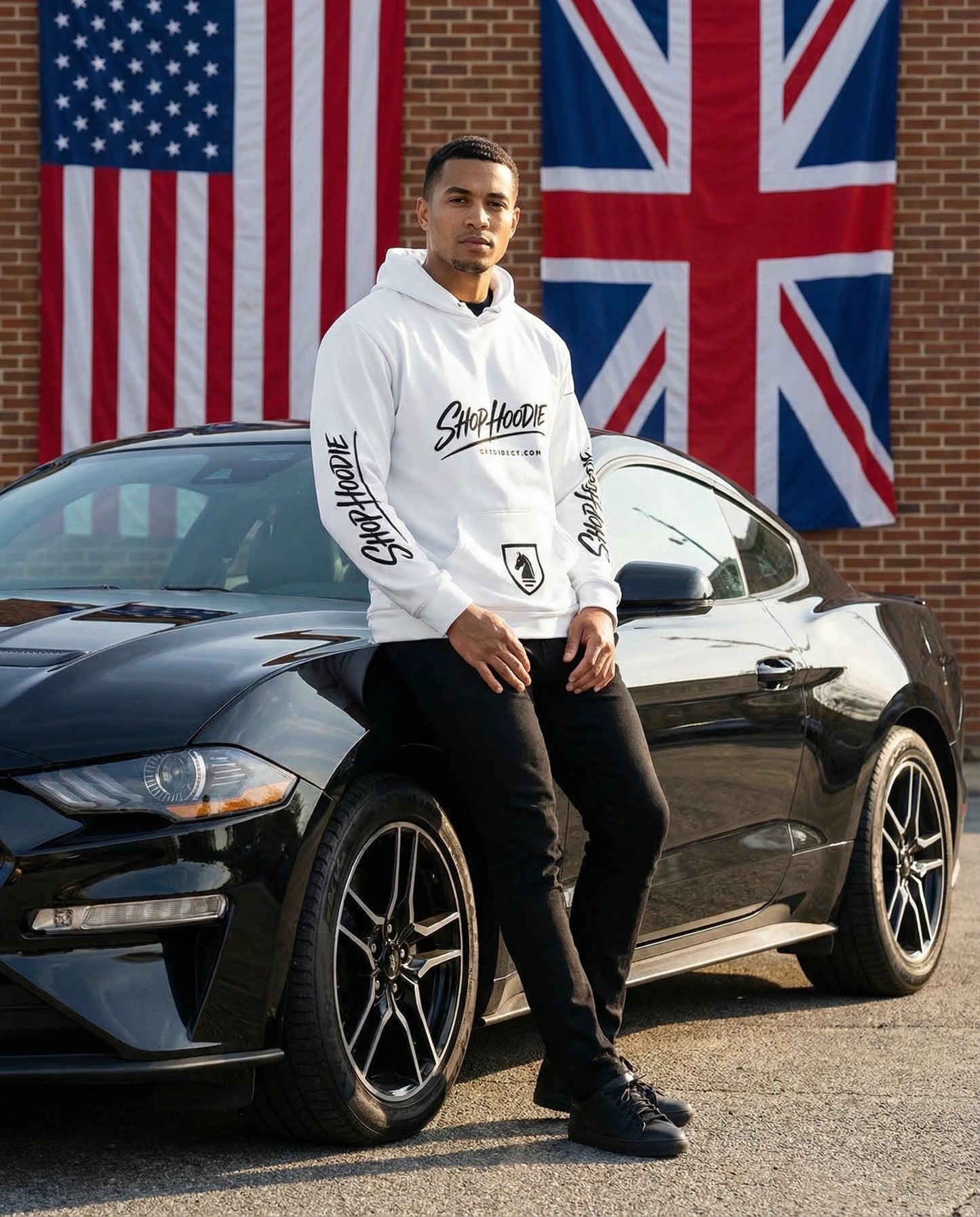 Person wearing a white hoodie with text, standing next to a black car with American and British flags in the background.