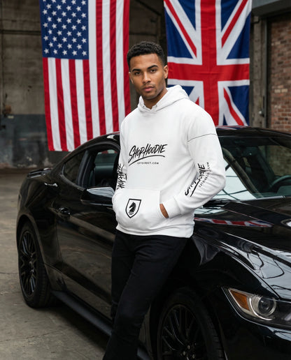 Man wearing a white hoodie with text and graphics, standing next to a black car with American and British flags in the background.
