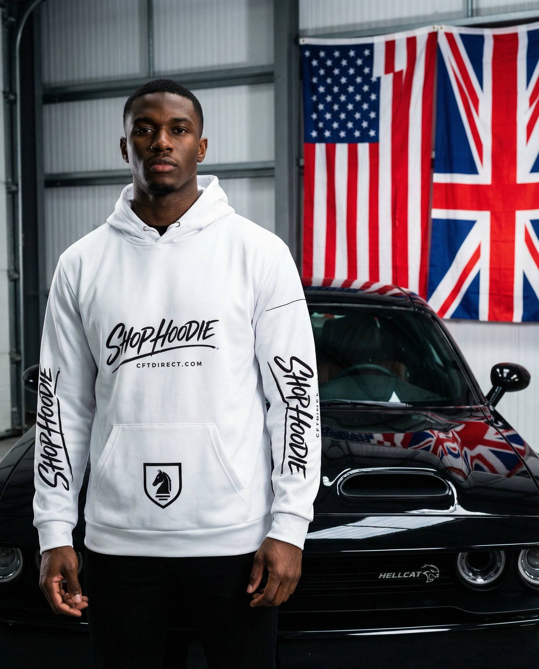 Person wearing a white hoodie with branding in front of a car and flags.