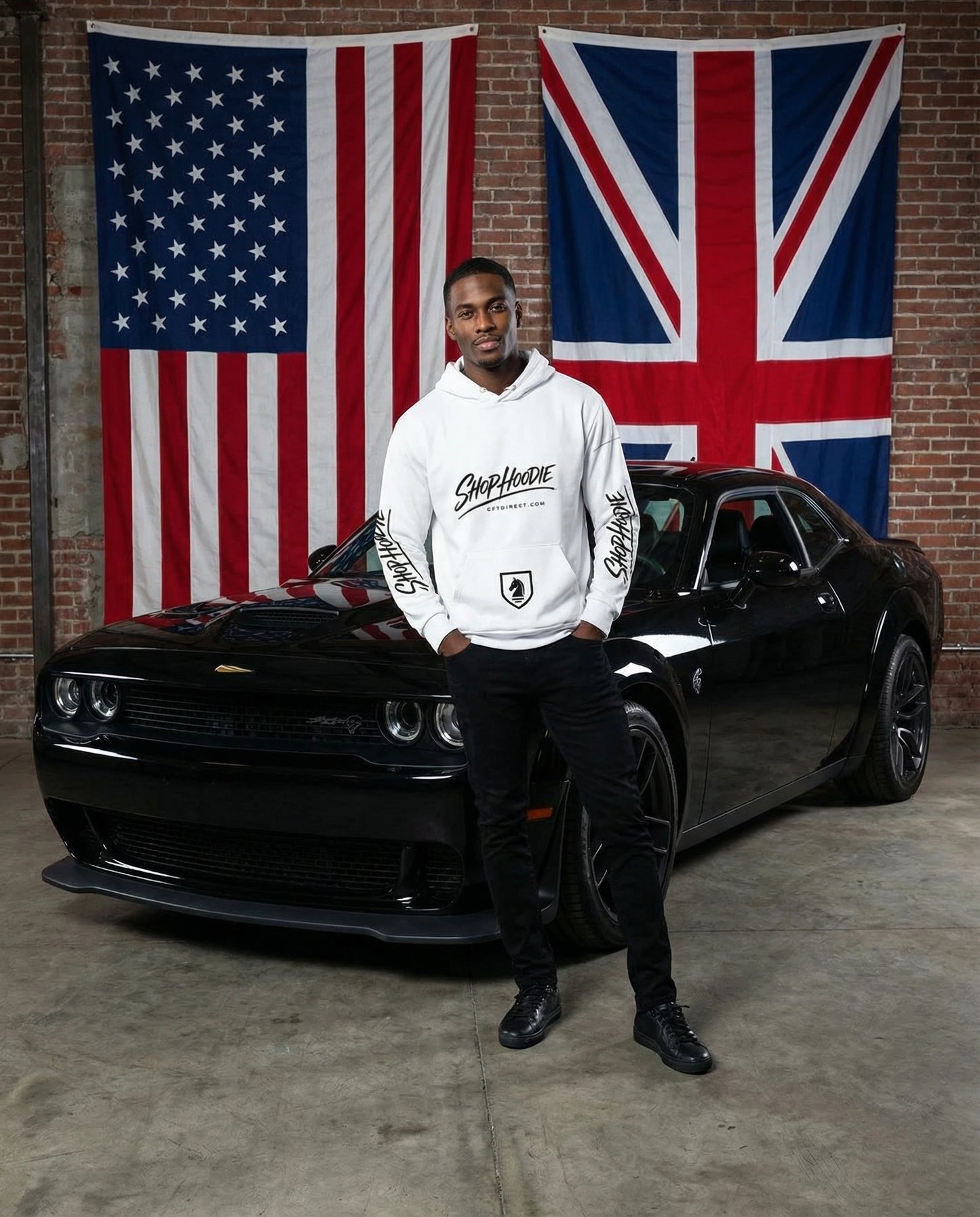 Man in a white hoodie standing next to a black car with American and British flags in the background.