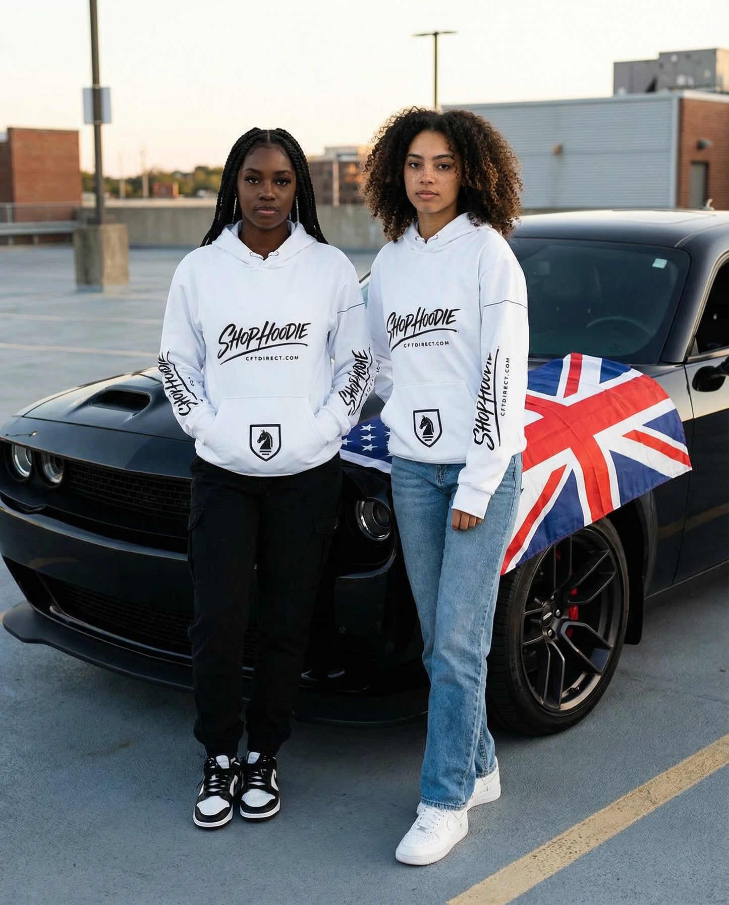 Two people wearing white hoodies with a brand logo, standing in front of a black car with a Union Jack flag.