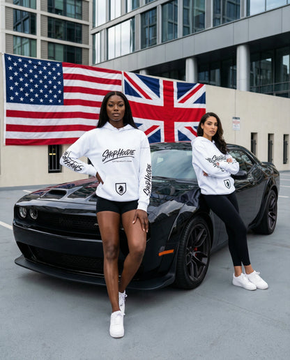 Two women in white sweatshirts with logos standing in front of a black car with American and British flags in the background.