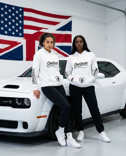 Two people wearing white hoodies with a logo in front of a white car and flags.