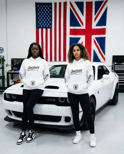 Two people wearing white hoodies with a logo in front of a white car and flags in the background