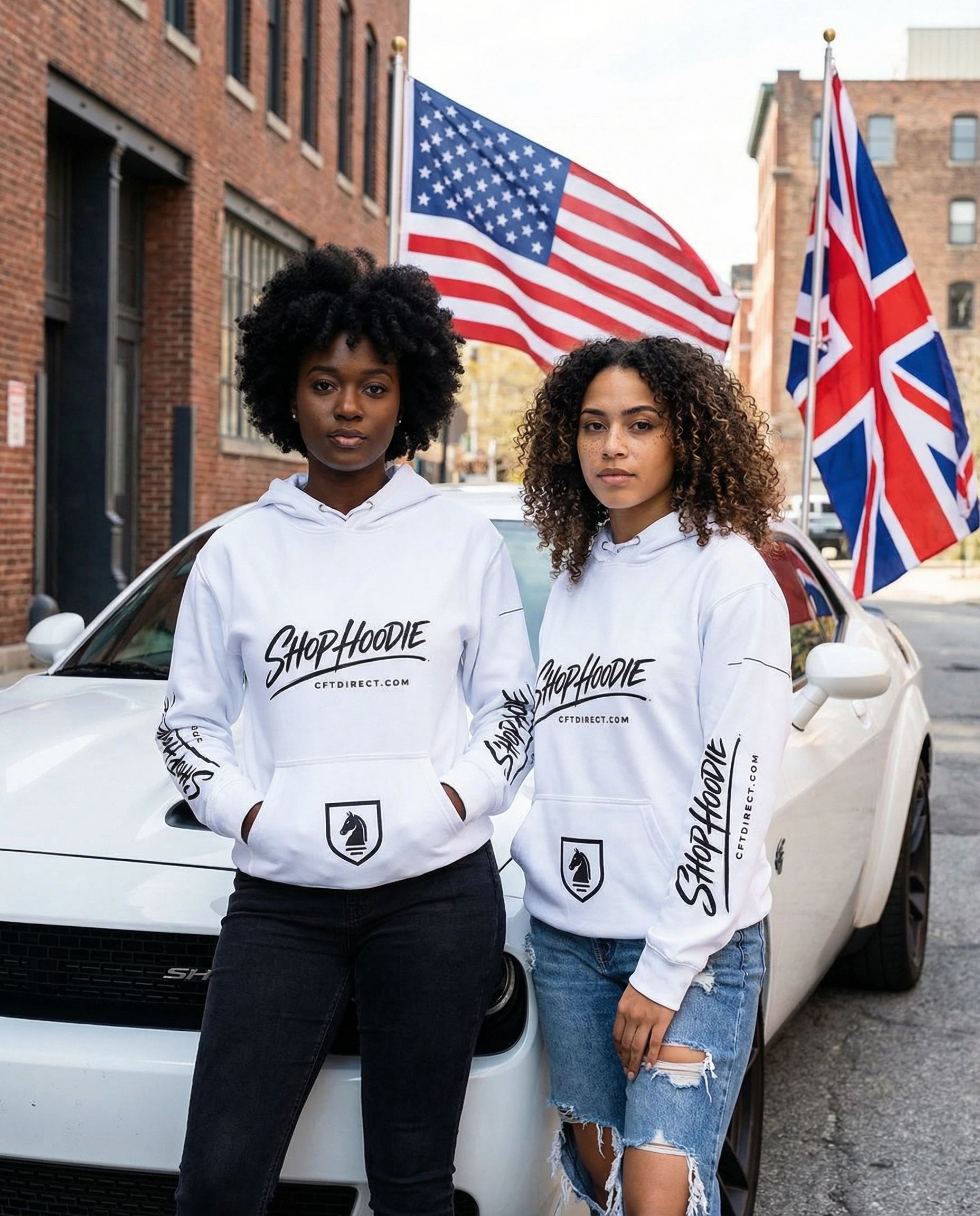 Two people wearing white hoodies with branding, standing in front of a white car with American and British flags in the background.