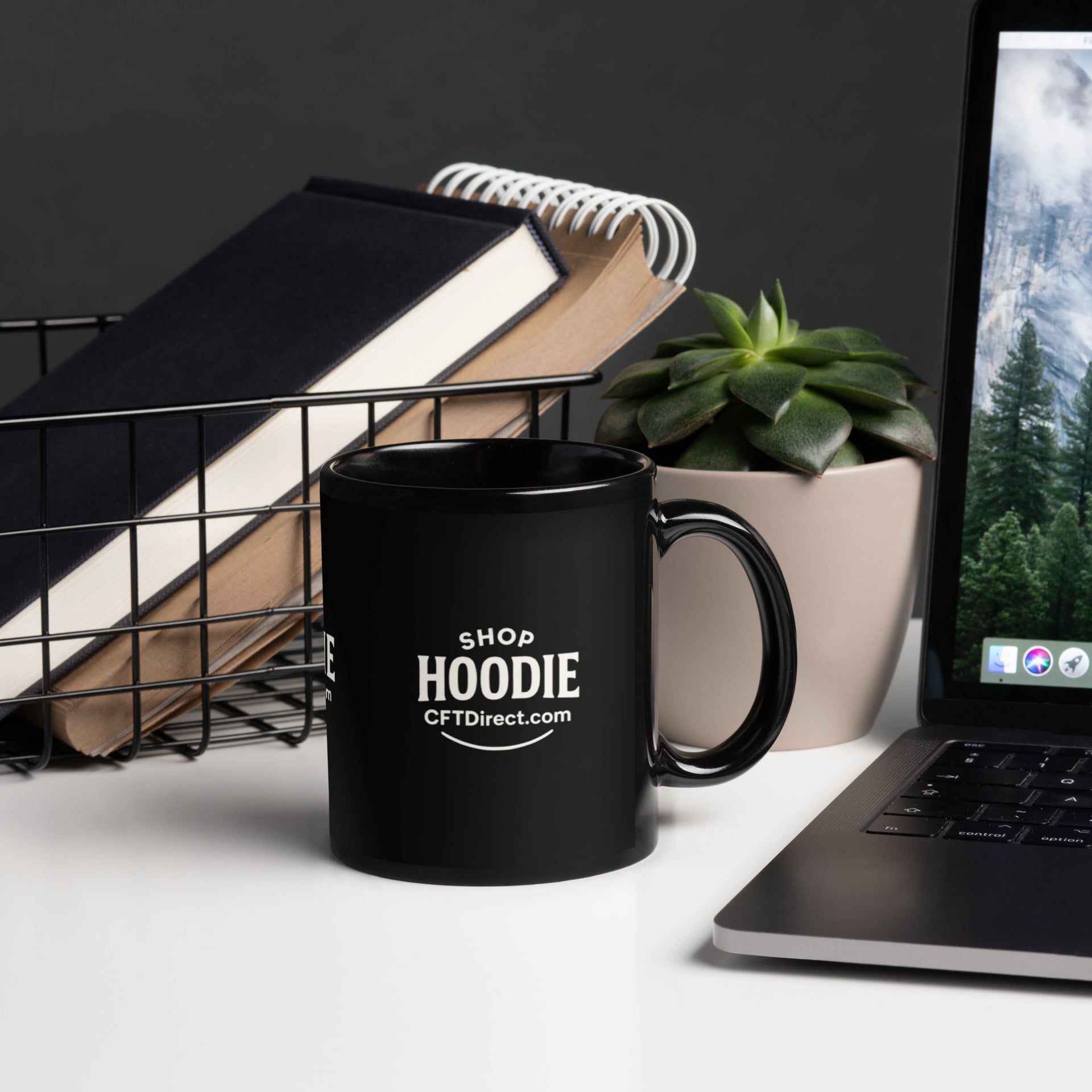 Black mug with 'ShopHoodie' branding on a desk with laptop, books, and plant.