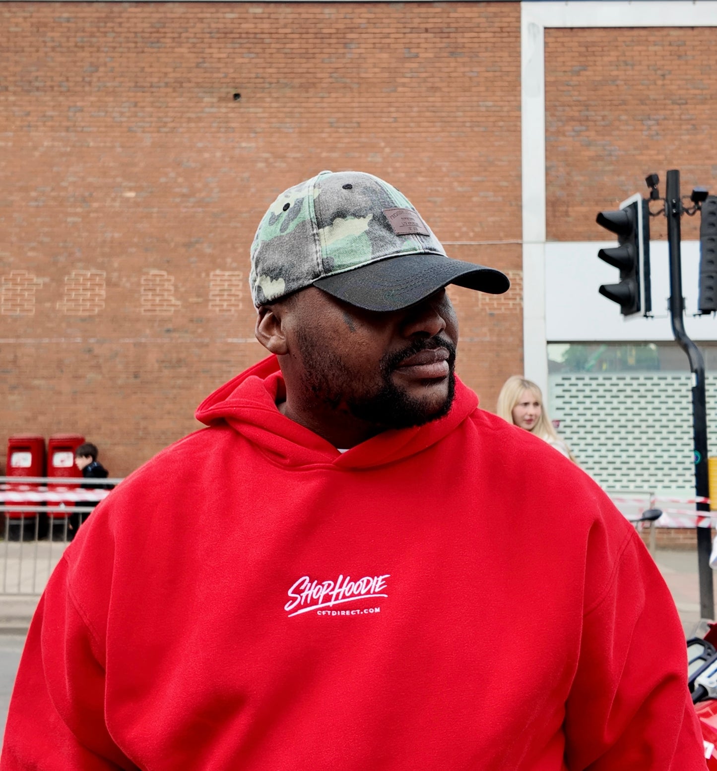Man wearing a red hoodie with a shophoodie brand logo, standing outdoors near a brick building.