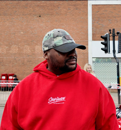 Man wearing a red hoodie with a shophoodie brand logo, standing outdoors near a brick building.