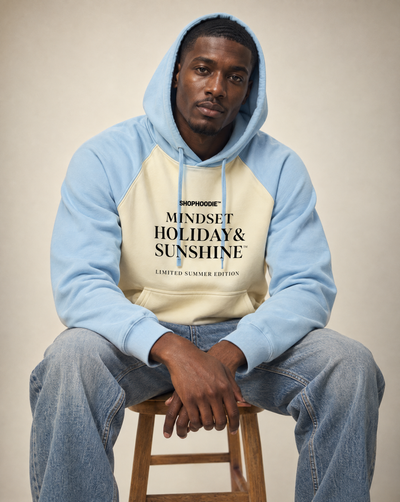 Male model seated wearing the SHOPHOODIE® Mindset Holiday & Sunshine Raglan Hoodie in Mild Apricot with Sky Blue, hood up